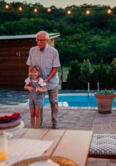 Smiling grandfather i playing with his grandchild in the backyard near pool, while his daughter setting dining table beside them
